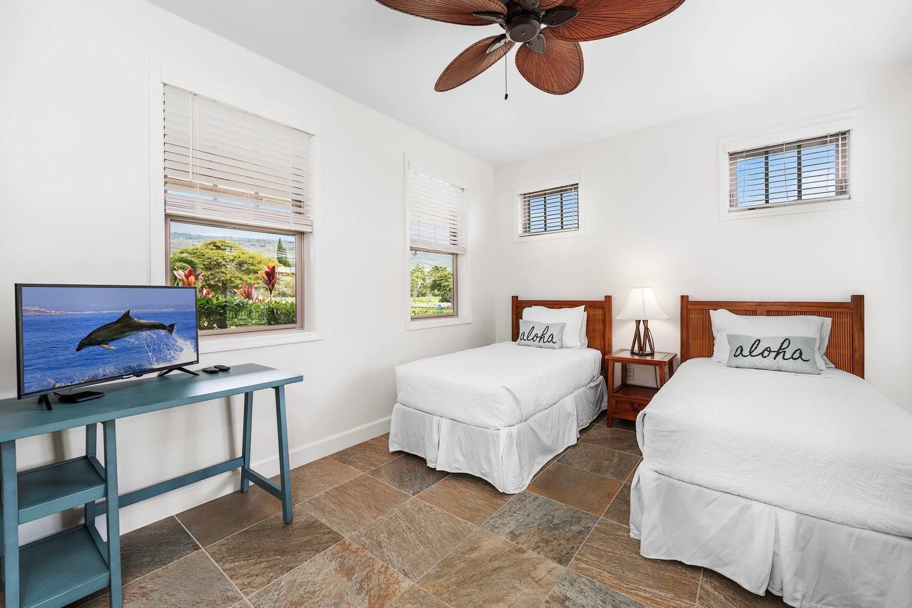 A bright bedroom with two beds, a television, and windows overlooking greenery.
