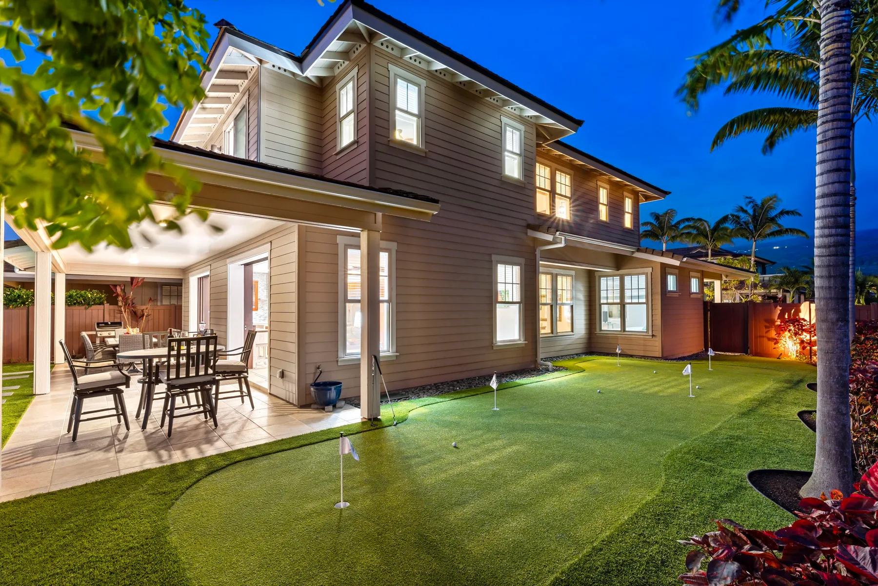 A large house at dusk overlooks a lawn, with a covered patio and outdoor seating.