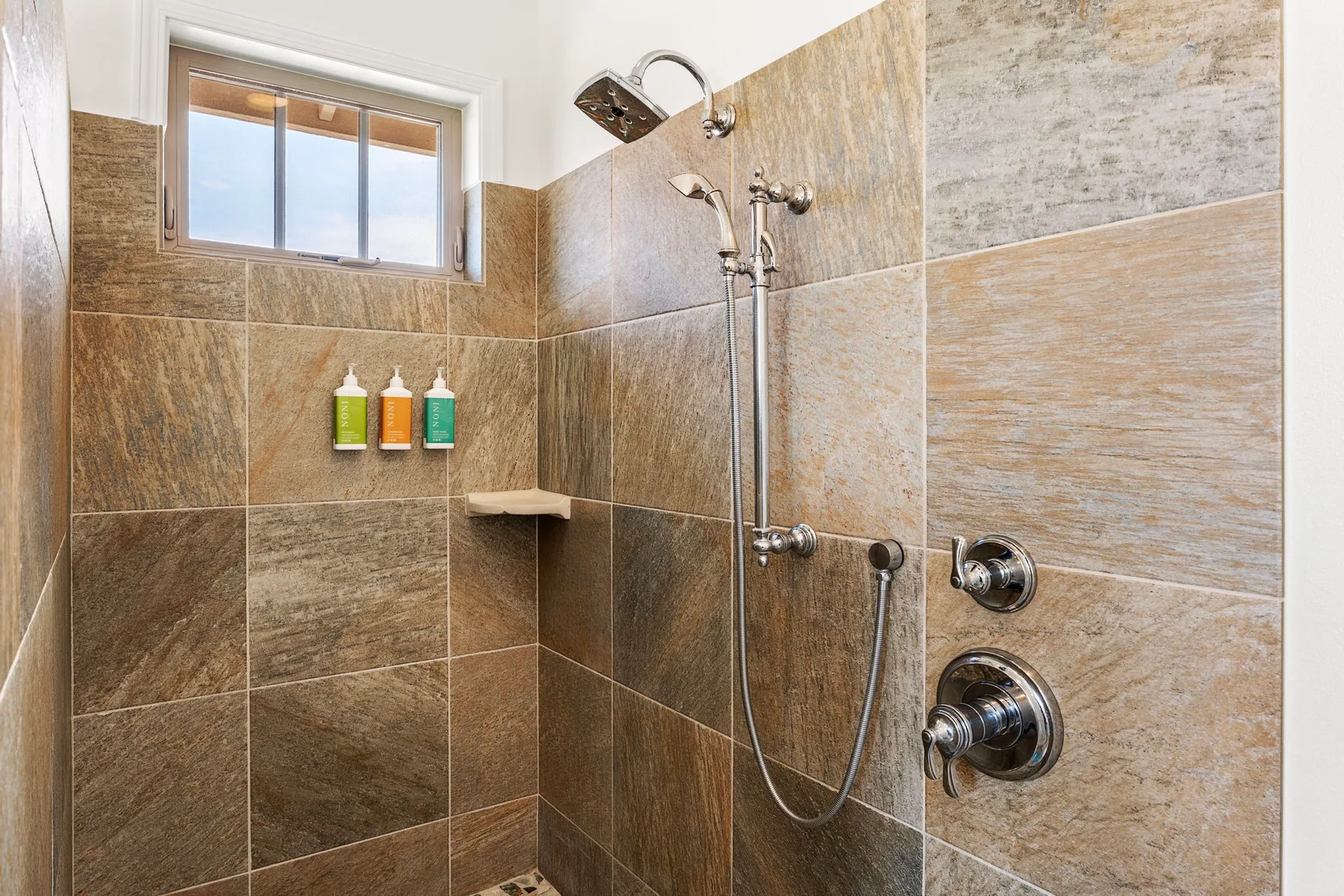 A tiled shower with a handheld showerhead and a small window.
