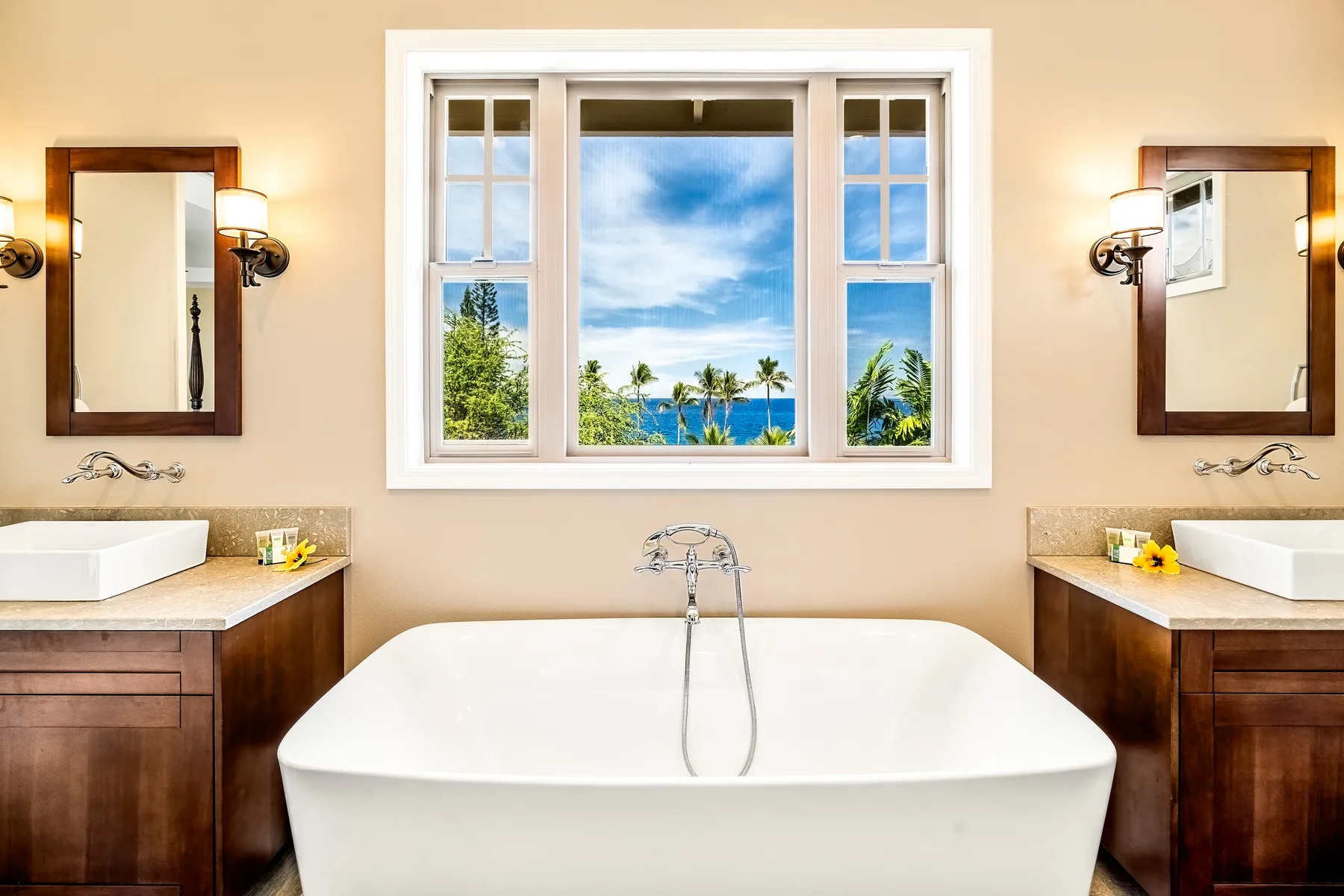 A bright bathroom features a freestanding bathtub beneath a large window overlooking palm trees and the sea.