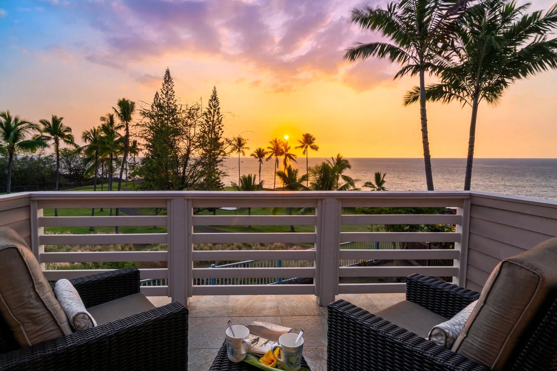 A balcony seating area overlooks palm trees and the ocean at sunset.