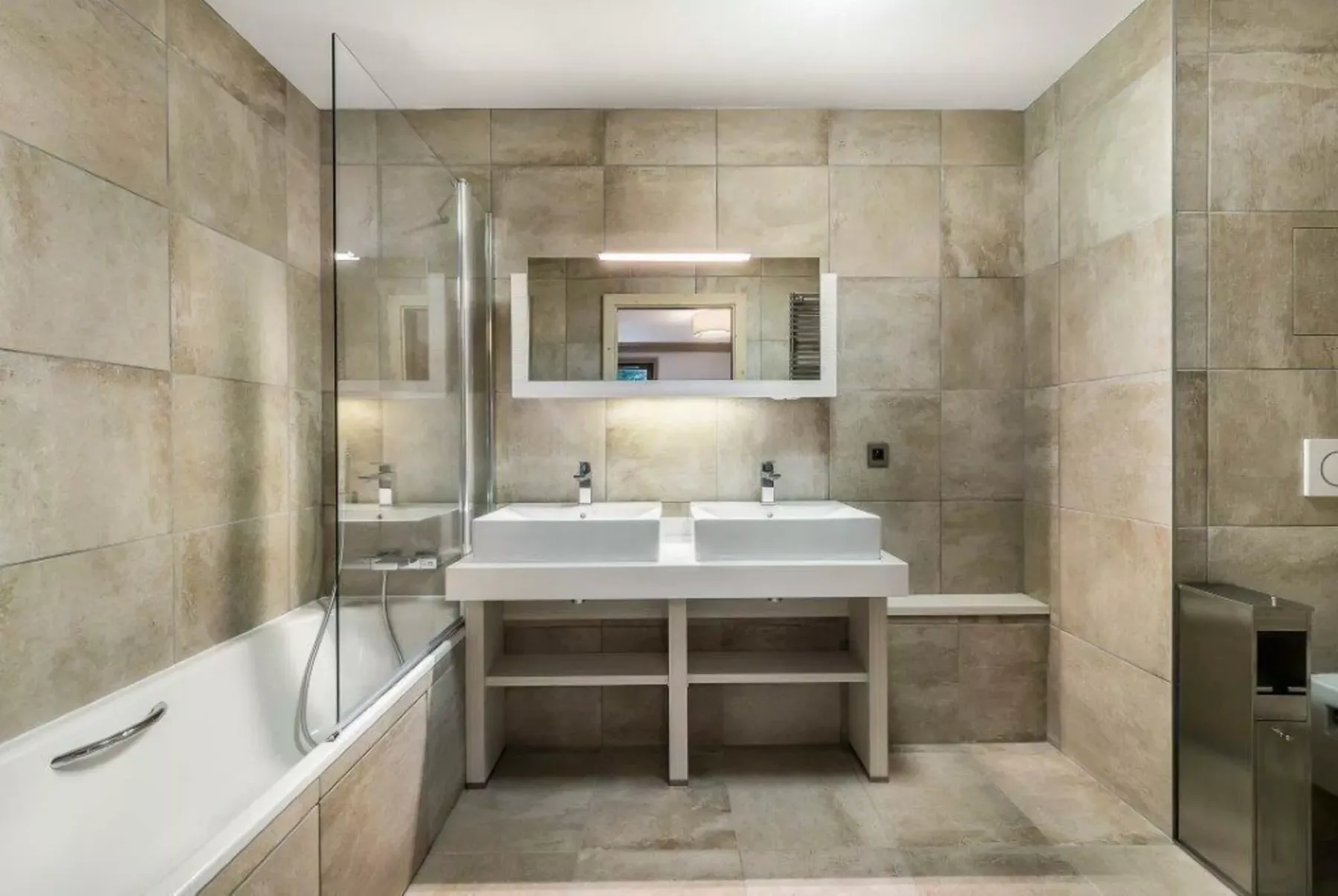 Un baño revestido de azulejos con bañera, mampara de ducha de vidrio y lavabo doble.