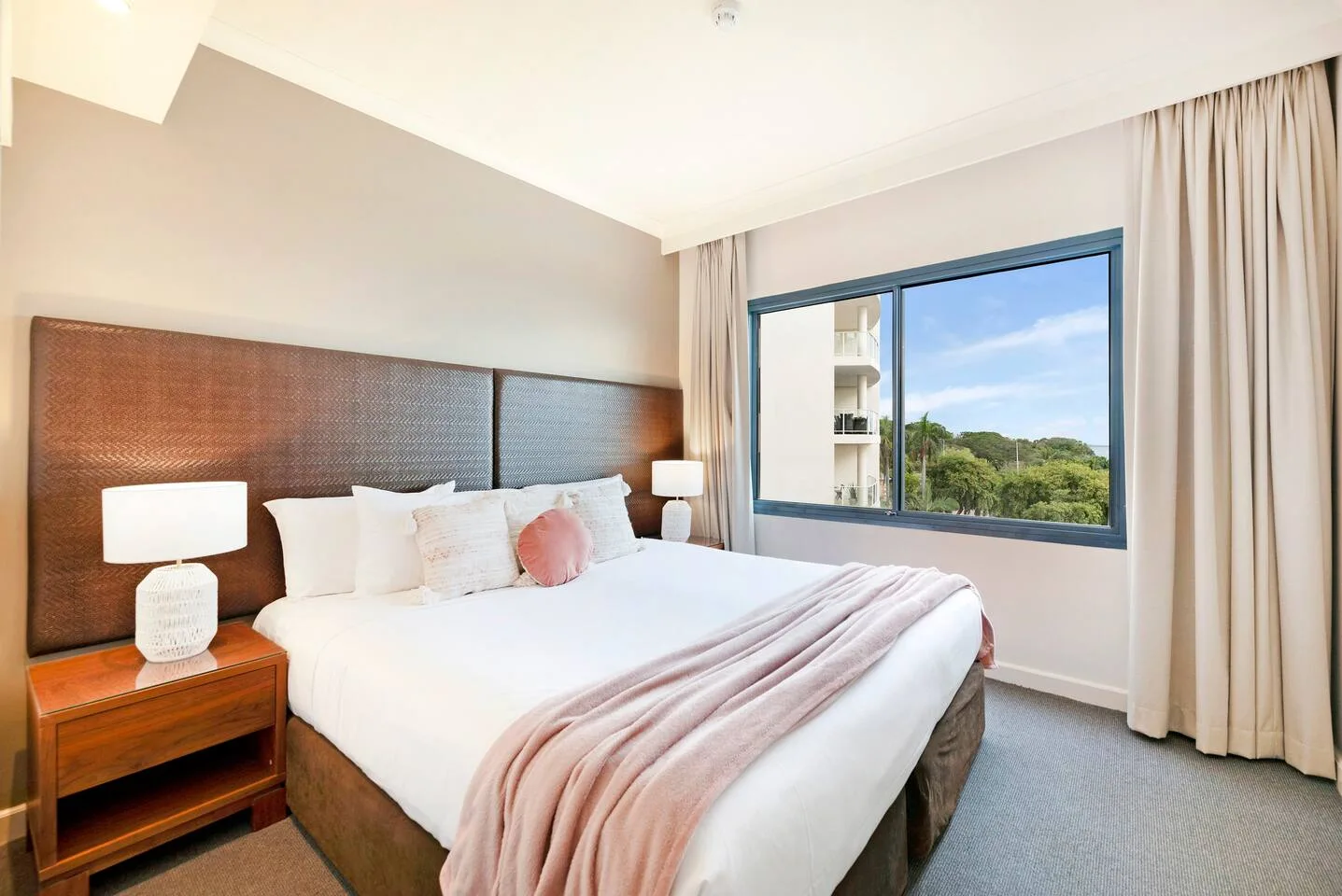 A bedroom with a large bed, bedside lamps, and a wide window overlooking greenery.