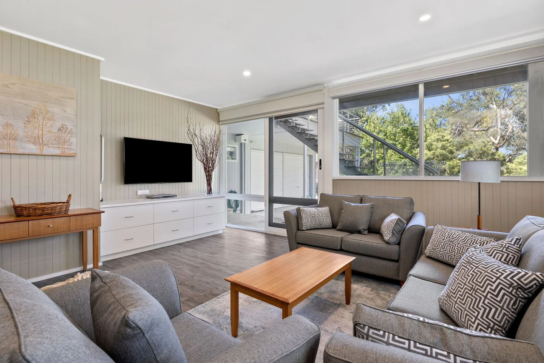 A bright living room with gray seating, a television, and large windows.