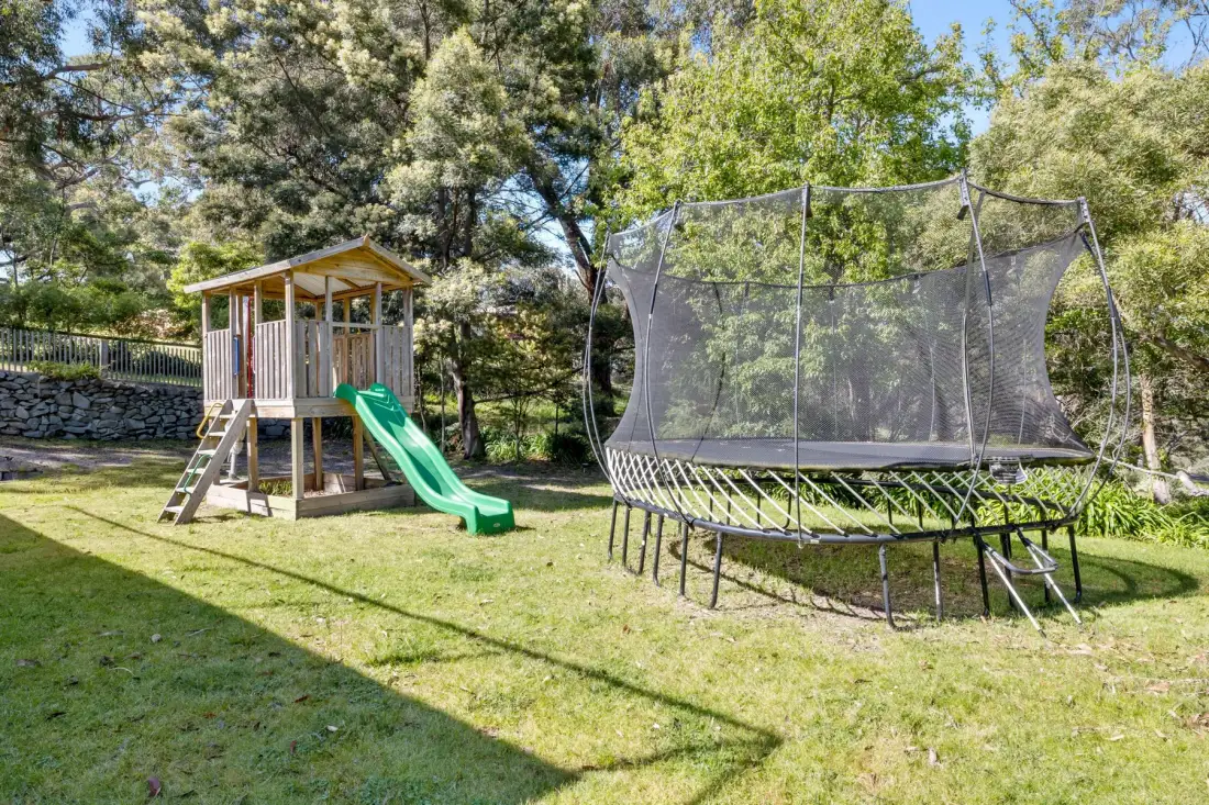 A grassy outdoor play area has a wooden play structure with a green slide and trampoline.