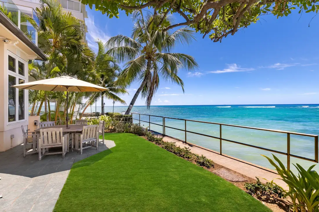 A seaside patio with outdoor seating overlooks the ocean beneath palm trees.
