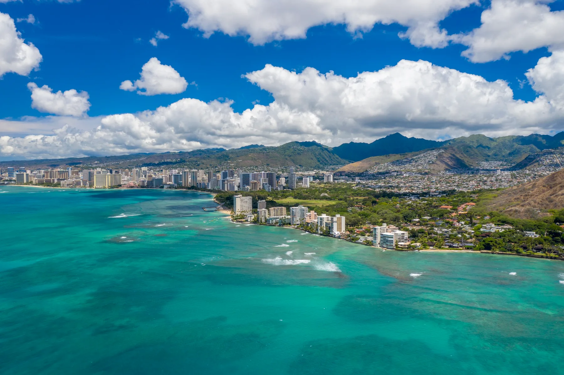 Aerial coastal cityscape with turquoise water, shoreline buildings, and mountains in the distance.