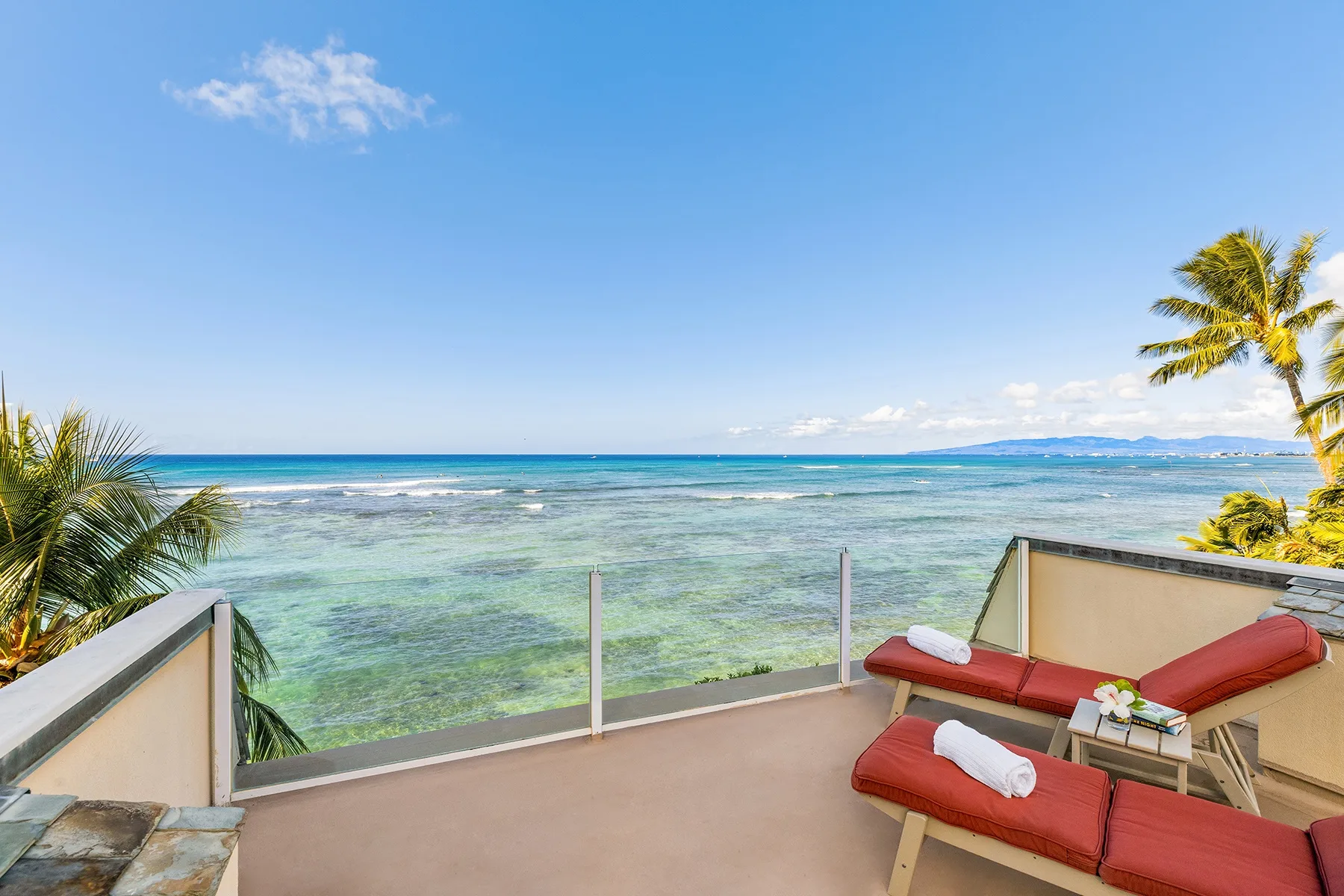 A seaside terrace overlooks clear water, with red lounge seating in the foreground.