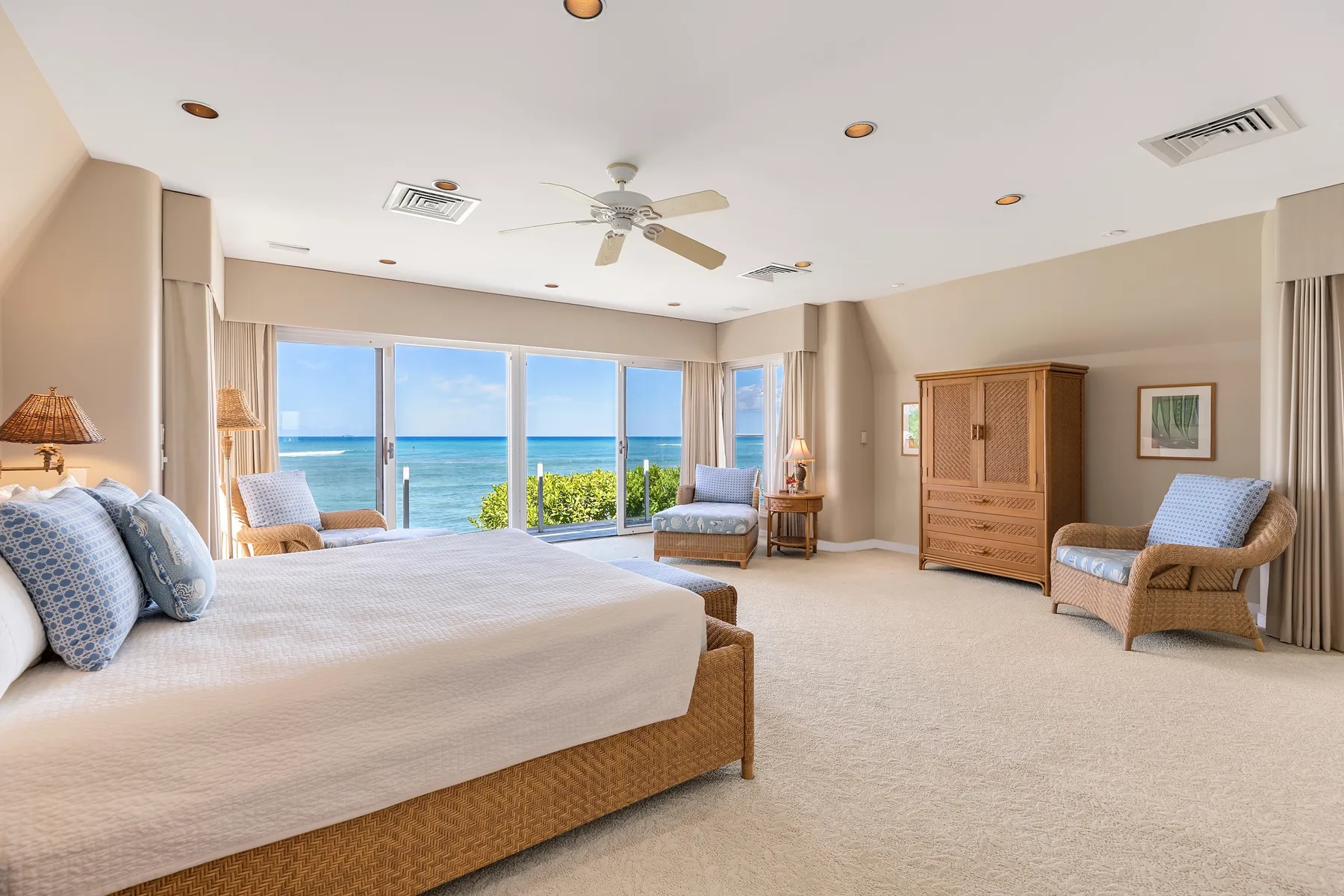 A bright bedroom with a bed, seating, and large windows overlooking the ocean.