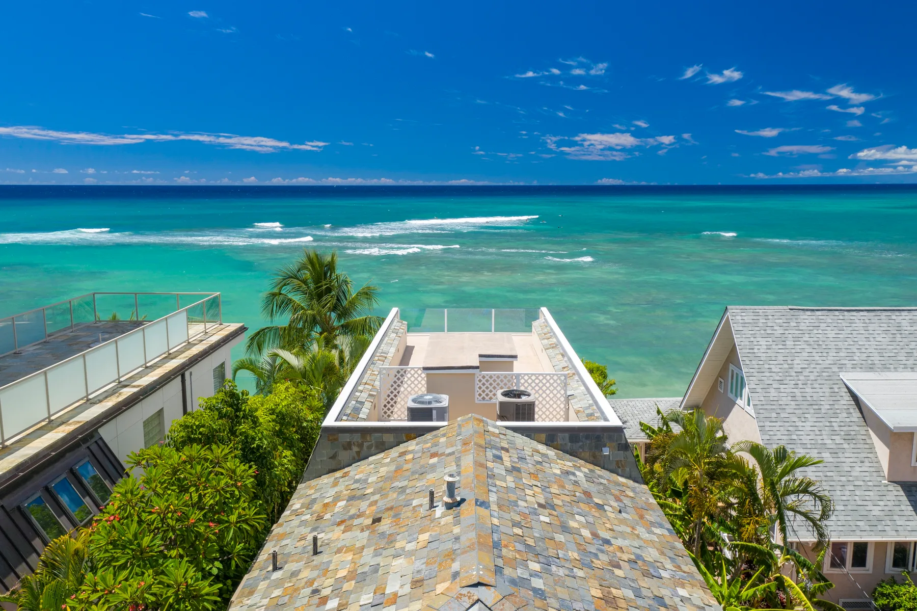 Aerial view of coastal buildings and palm trees beside turquoise ocean water.