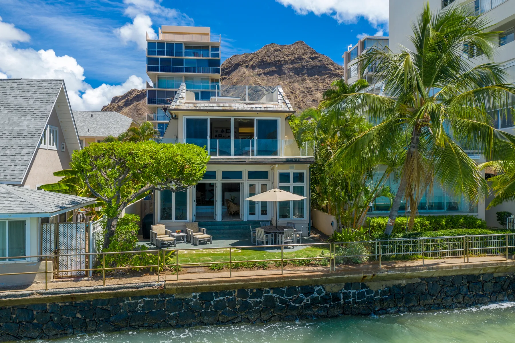 A modern waterfront building with glass windows, palm trees, and a mountain backdrop.