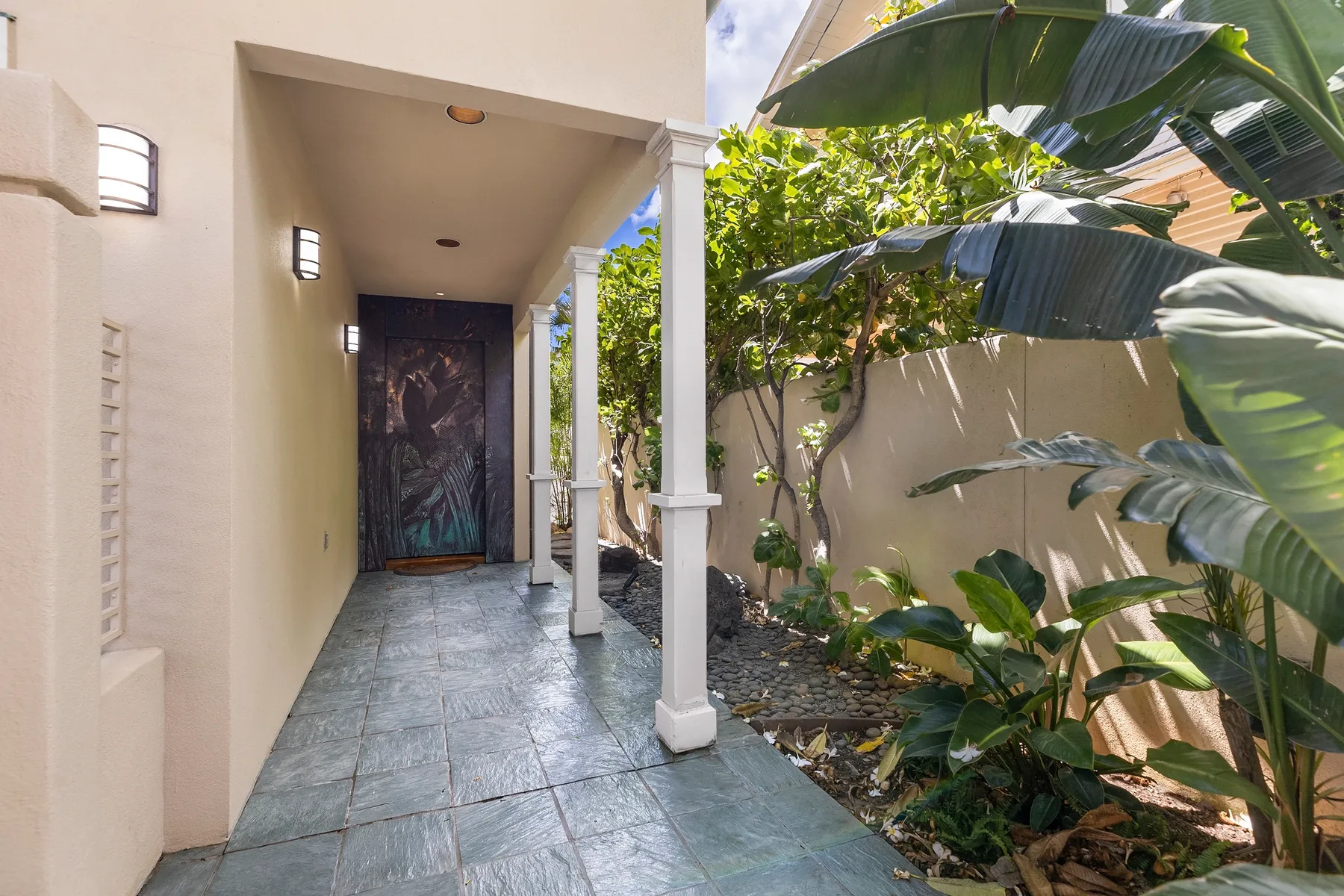 A covered outdoor walkway with tiled flooring, columns, and leafy plants.