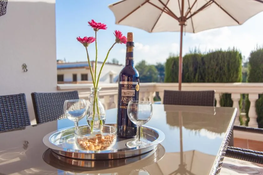 Comedor al aire libre con mesa de vidrio, copas de vino y una sombrilla.