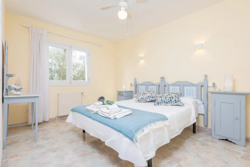 Un dormitorio luminoso con una colcha azul, muebles auxiliares y una ventana.