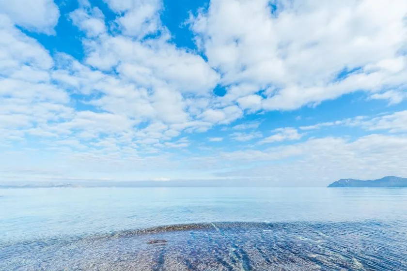 Un paisaje marino tranquilo se extiende bajo un cielo azul nublado, con una costa distante.