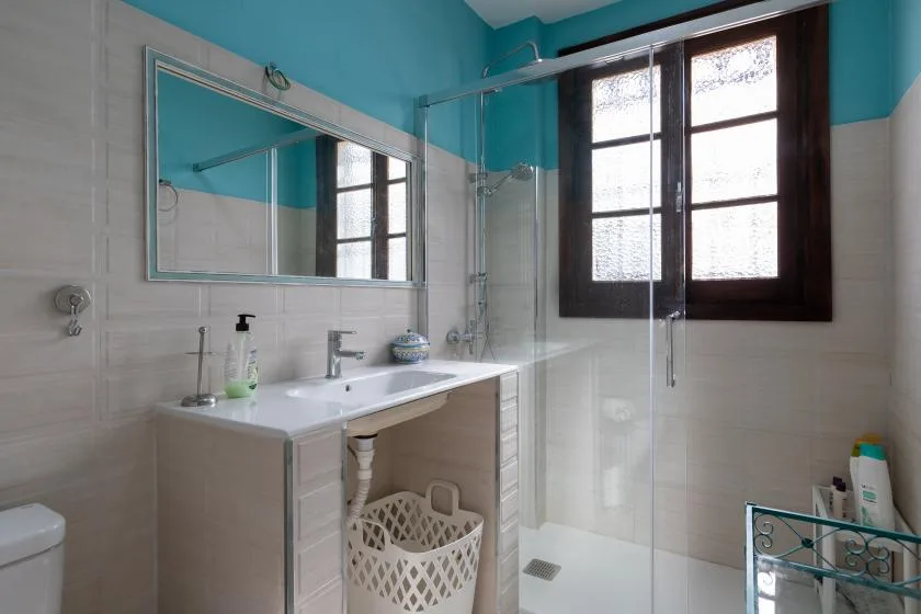 A bathroom with a sink, mirror, and glass shower beside a window.