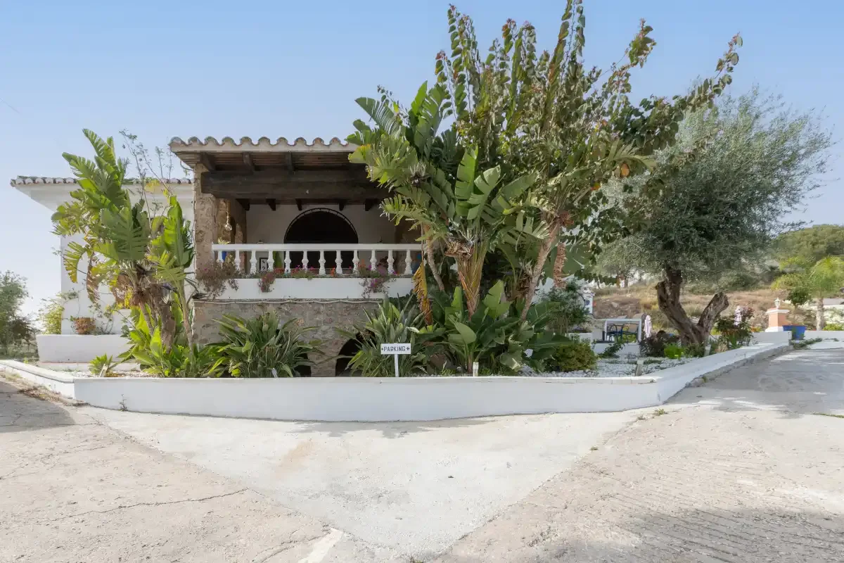 A white house with a shaded balcony stands among leafy plants beside a paved road.