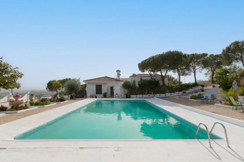 A rectangular outdoor swimming pool sits beside a low white building and trees.