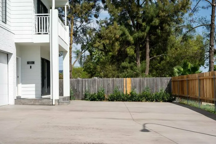 A paved outdoor area beside a white building, with trees and a wooden fence.