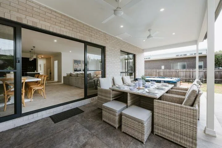 A covered outdoor dining area with a table and cushioned seating beside the house.
