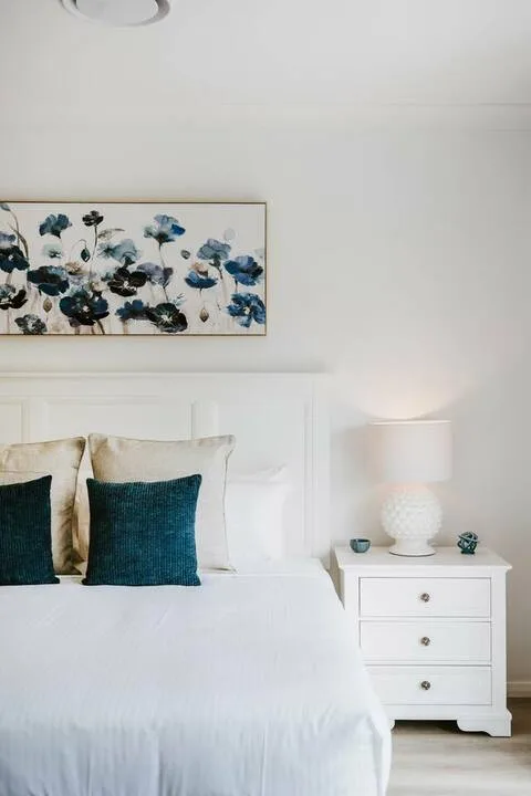 Bedroom with a white bed, dark accent pillows, and floral artwork above the headboard.