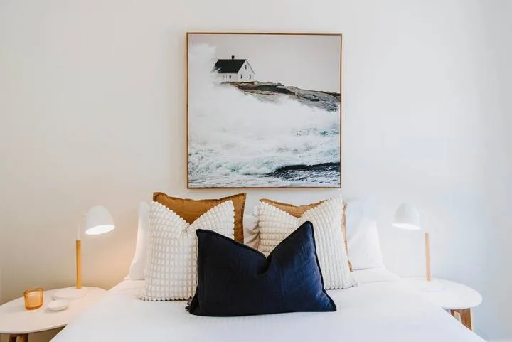 A bed with layered pillows sits beneath a framed coastal painting.