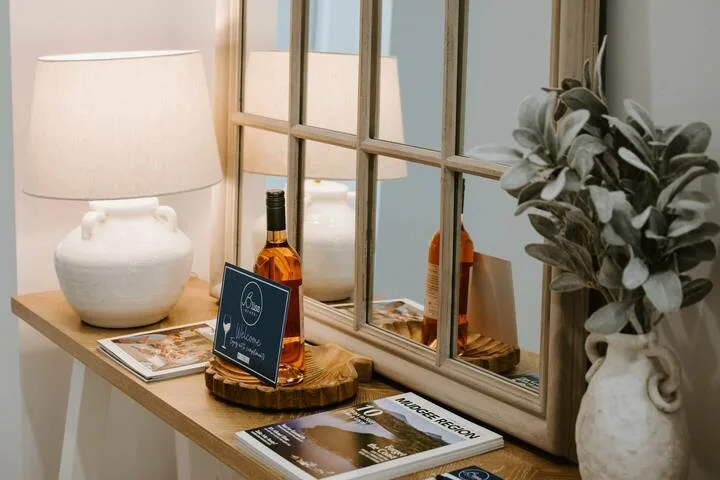 A wooden console displays a lamp, bottle, magazines, and a potted plant beside a framed mirror.
