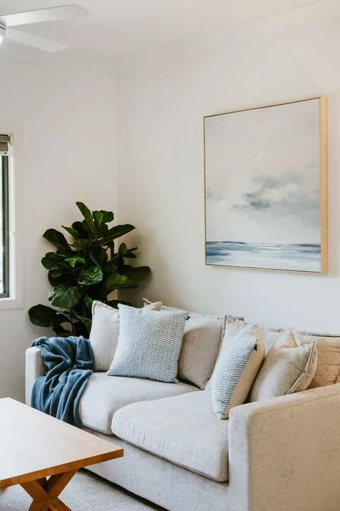 A bright living room with a light-colored sofa, leafy plant, and framed artwork.