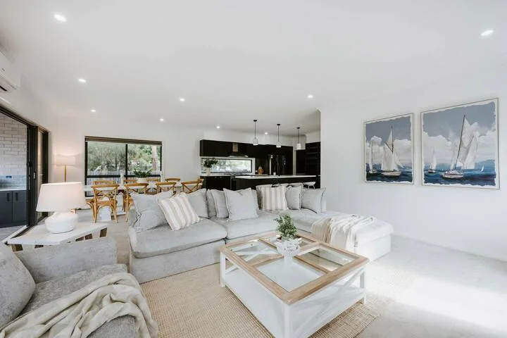 A bright living room with seating, a glass coffee table, and an open kitchen.