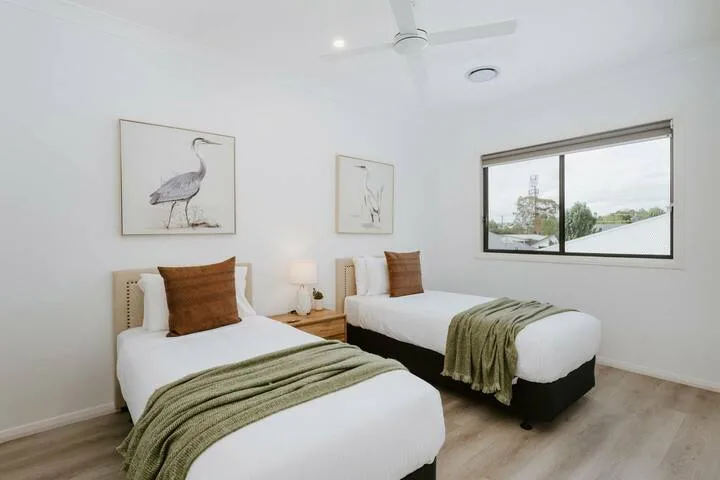 A bright bedroom with two beds, neutral bedding, and a large window.