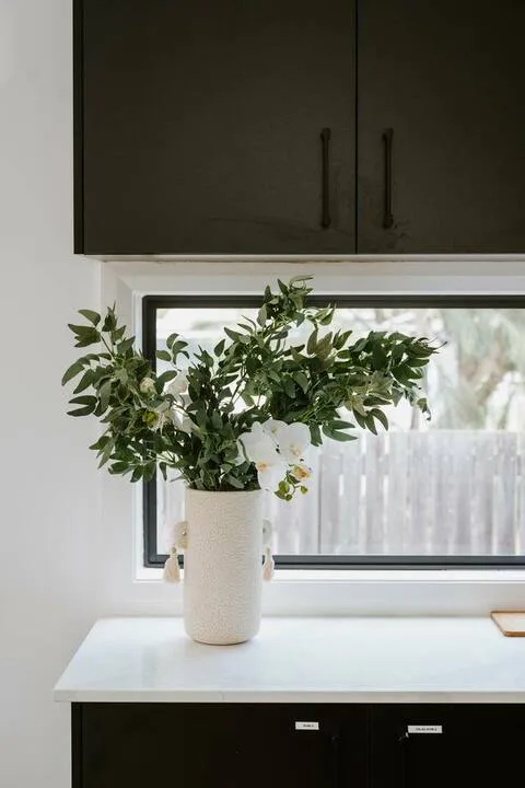 A kitchen counter holds a vase of leafy branches beneath dark cabinets.