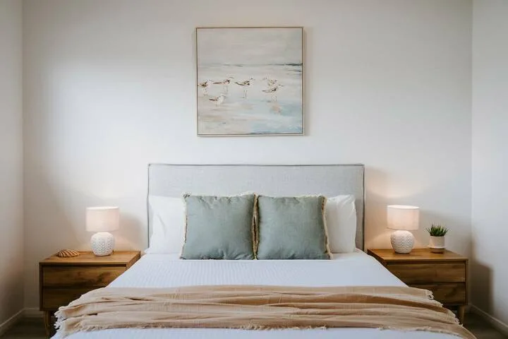 A bedroom with a neatly made bed, bedside tables, and coastal artwork.