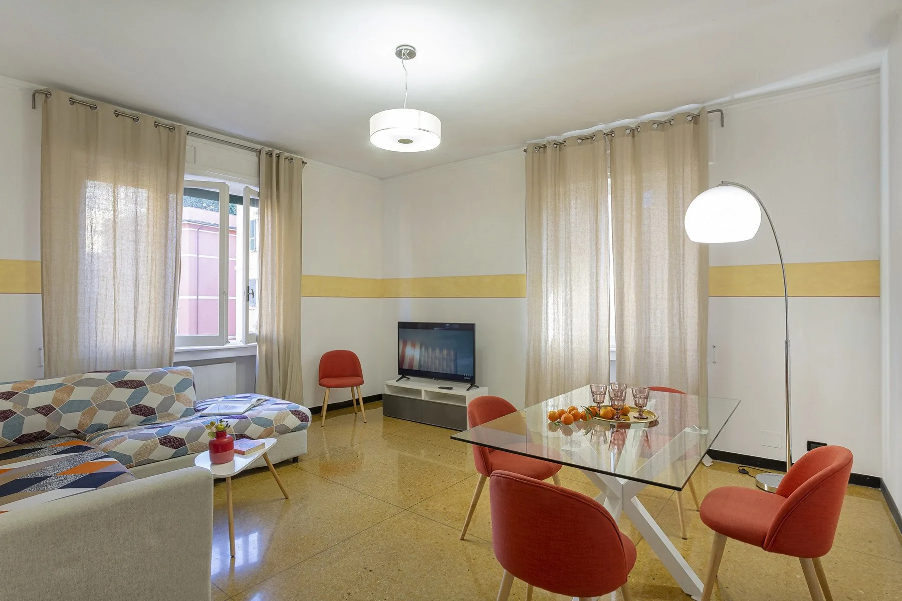 A living and dining room with patterned seating, a glass table, and curtained windows.