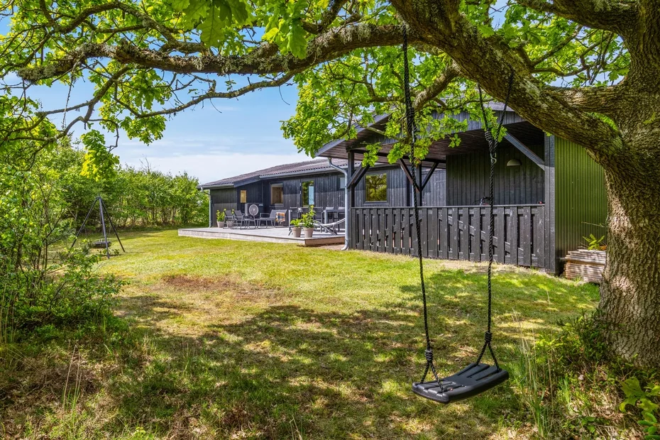 Una casa de fachada oscura junto a un patio de césped, bajo un árbol frondoso.