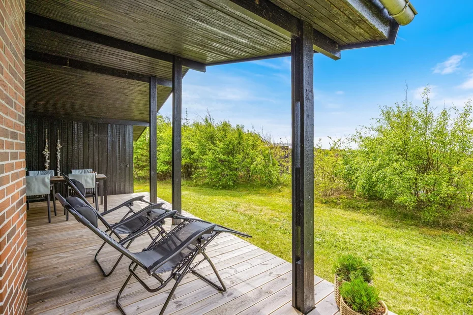 Una terraza cubierta de madera con asientos reclinables da a un jardín con césped y árboles.