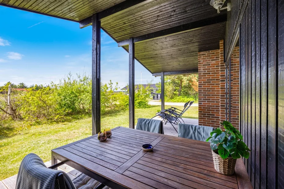Un patio exterior cubierto tiene una mesa de madera, asientos y vistas al jardín.