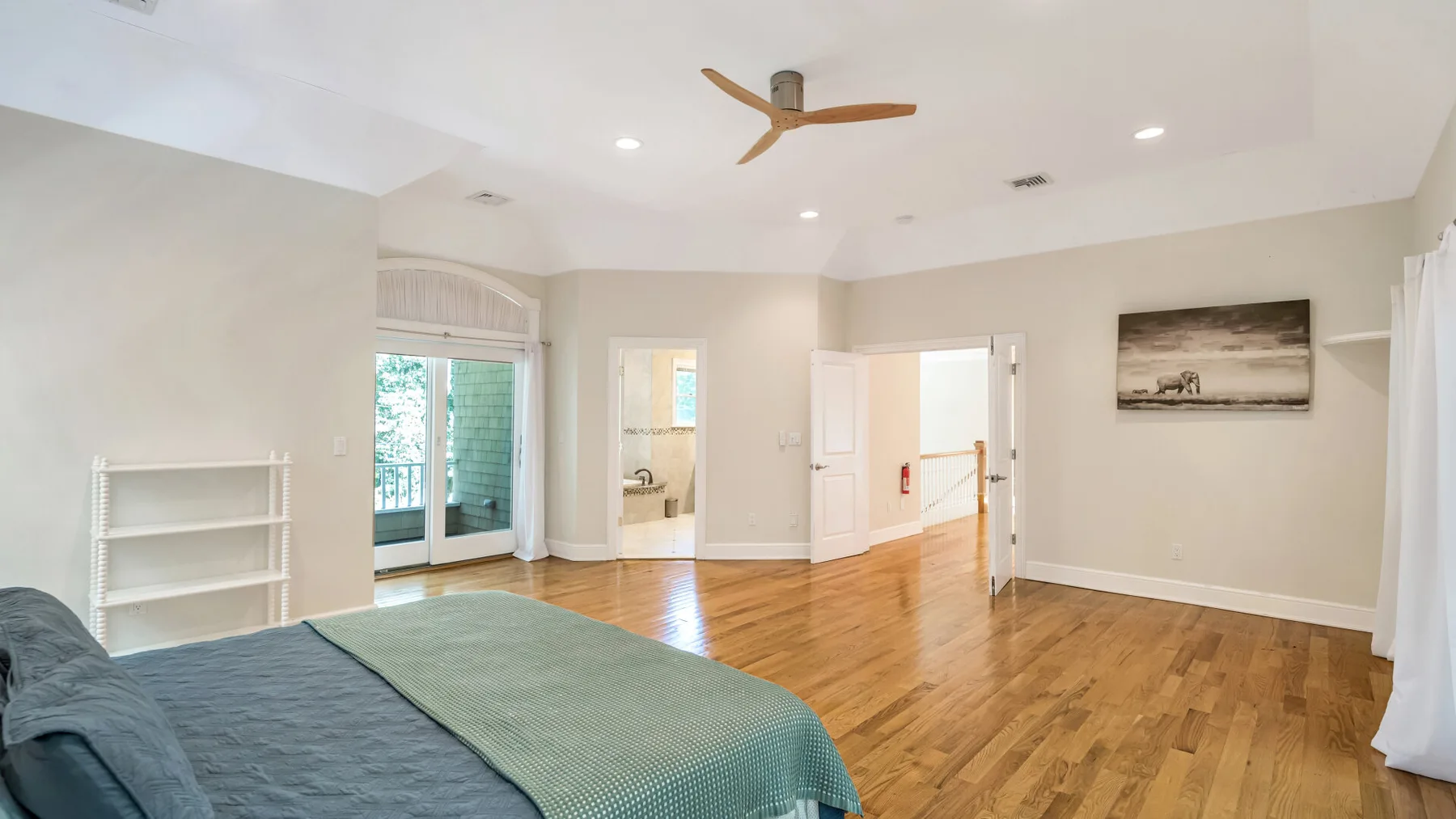 A bright bedroom with a bed, wood flooring, and doors opening to a hallway.