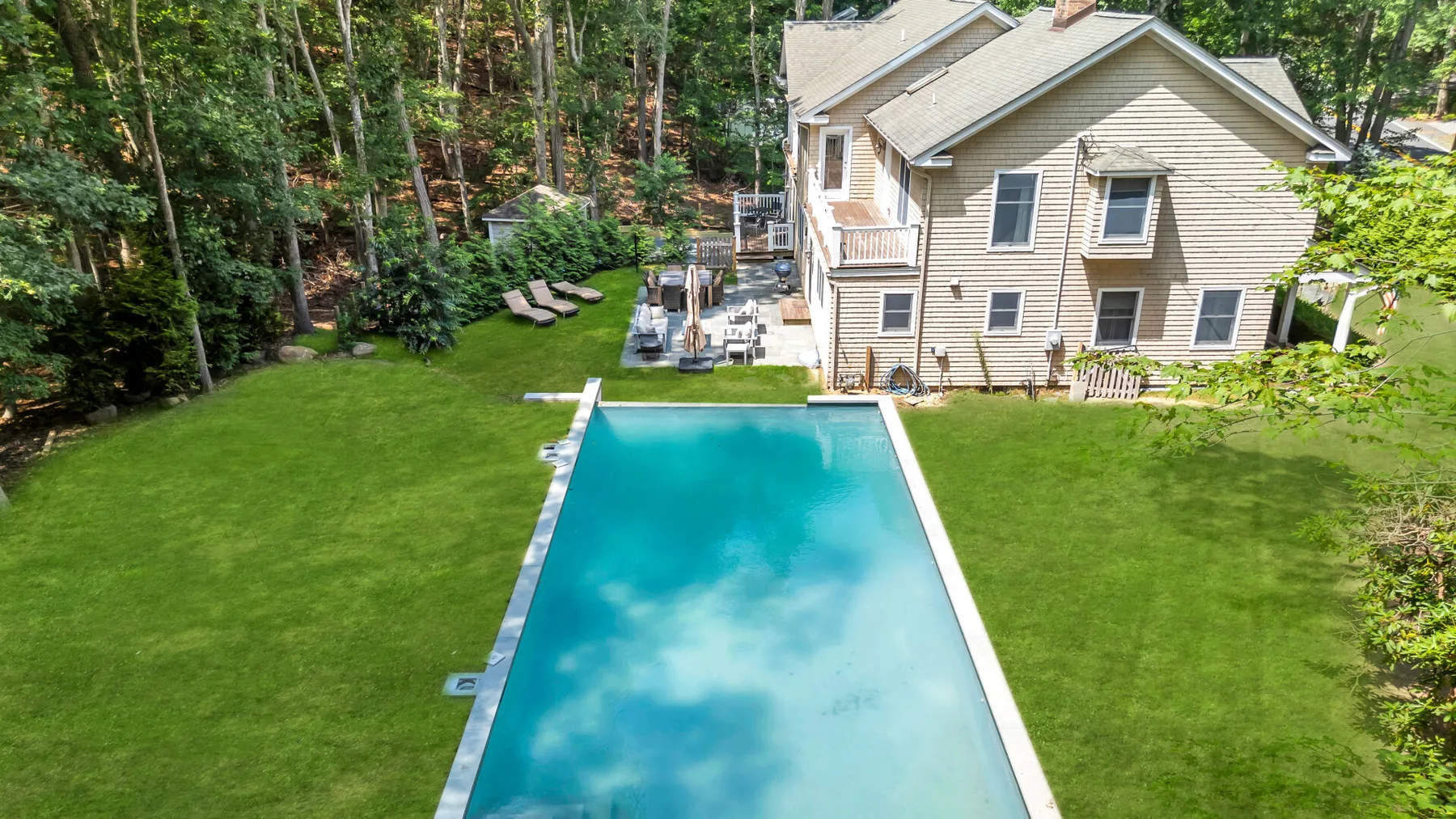 A swimming pool beside a house, surrounded by a green lawn and trees.