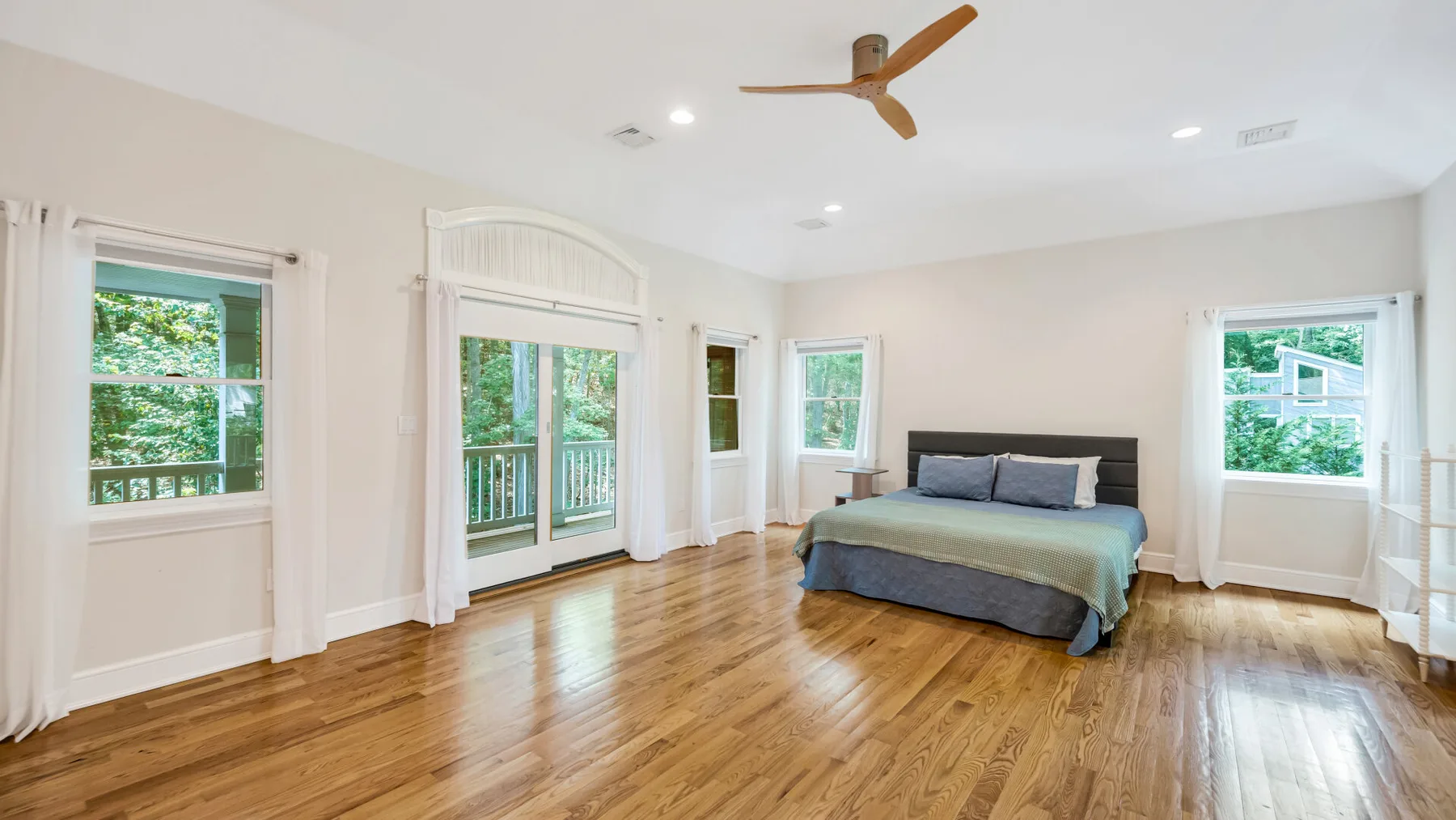 A bright bedroom with a bed, wooden flooring, and several large windows.