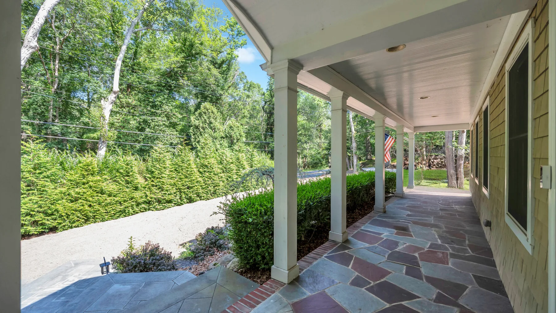 A covered porch with stone paving and green shrubs along the driveway.