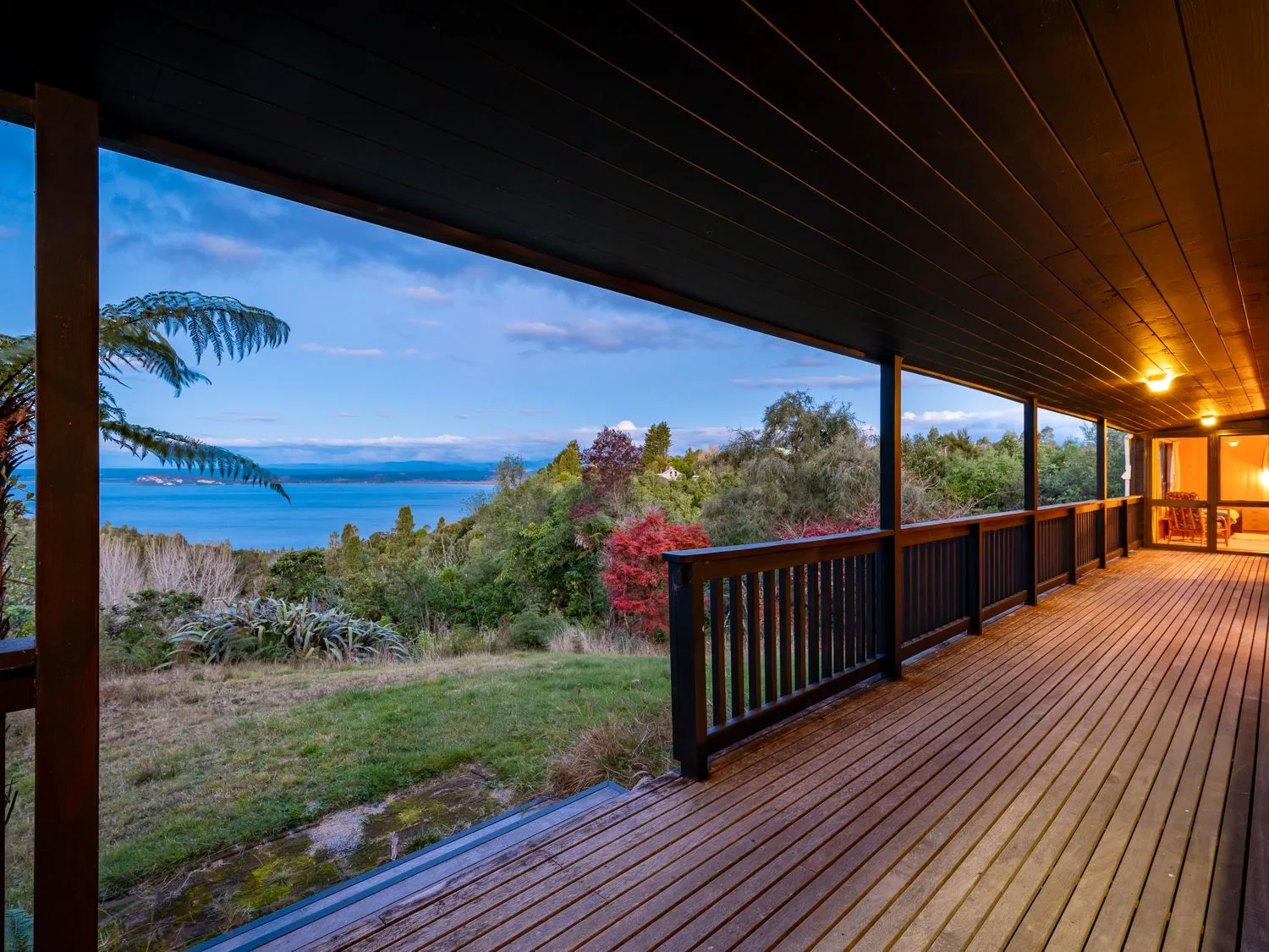 A covered wooden deck with railing overlooking trees and a lake at Lake View Lodge Taupo Holiday Home in Pukawa Bay
