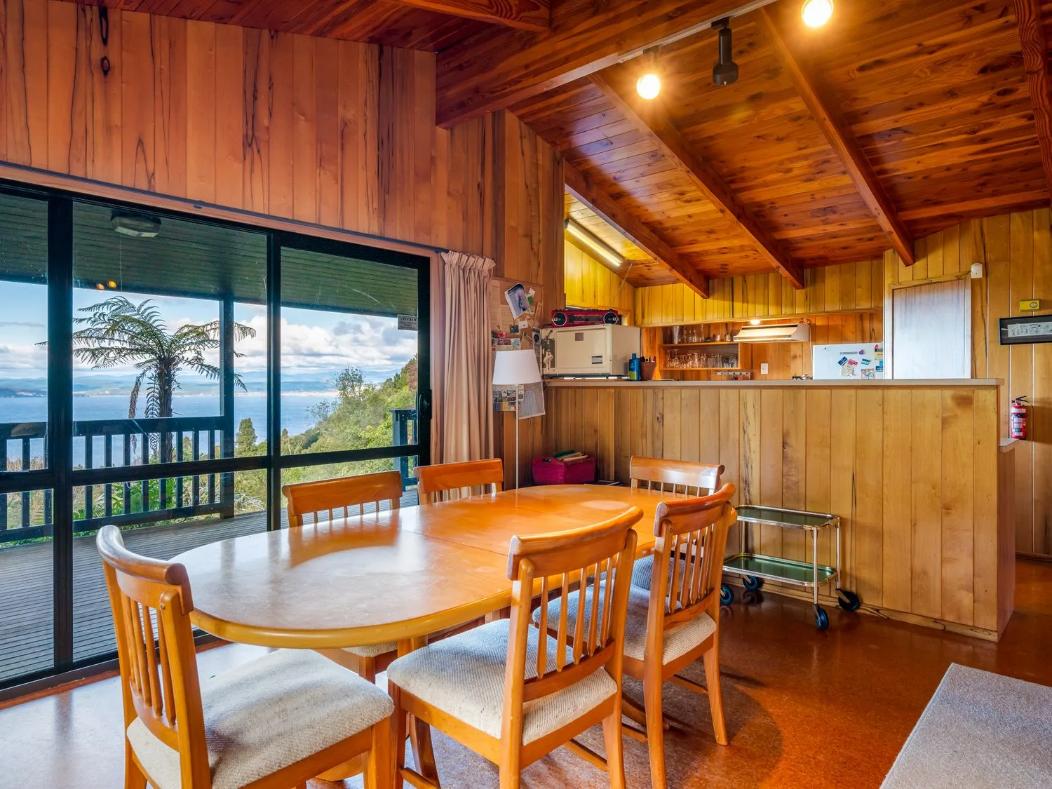 A dining area with wooden table and chairs next to sliding glass doors showing a view of trees and water at Lake View Lodge Taupo Holiday Home in Pukawa Bay