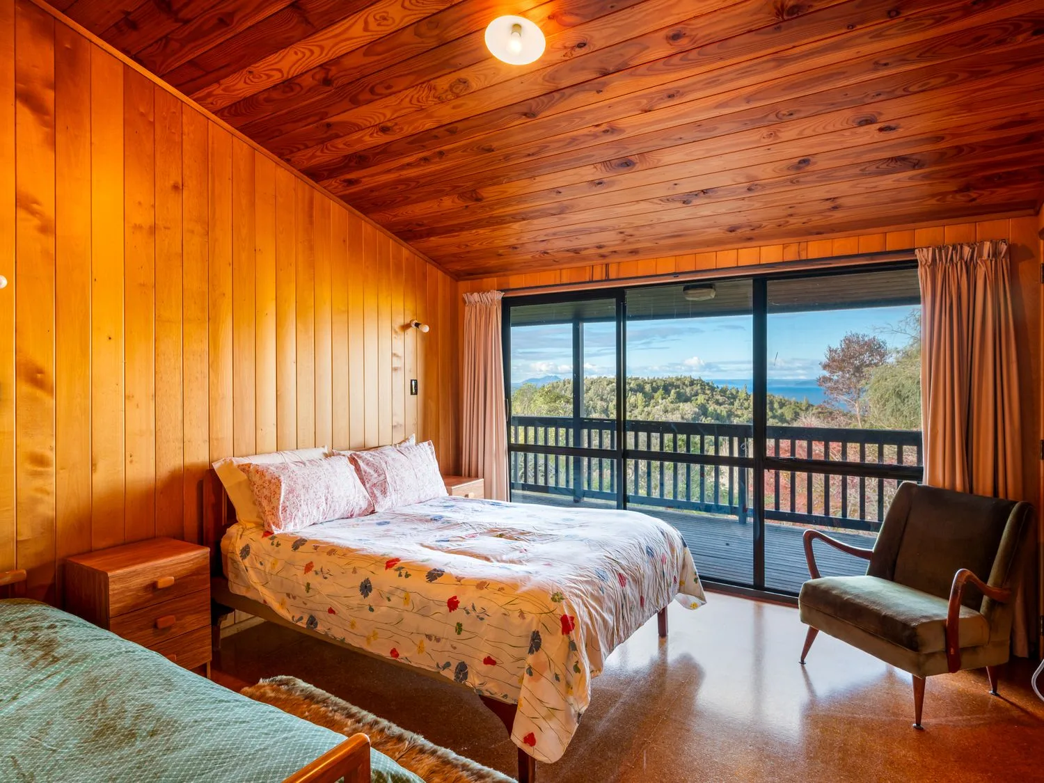 A bedroom with two beds a chair wooden walls and a sliding glass door overlooking a balcony and trees at Lake View Lodge - Taupo Holiday Home in Pukawa Bay