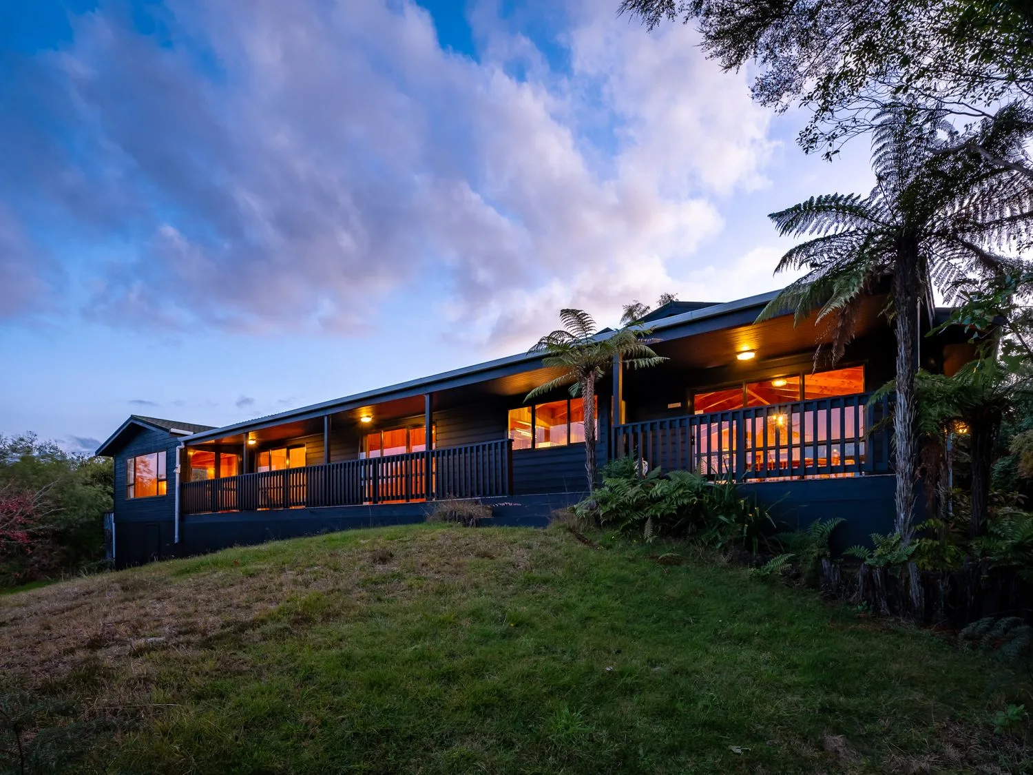 An exterior view of a single story house with a covered porch and lights on surrounded by trees and grass at Lake View Lodge Taupo Holiday Home in Pukawa Bay