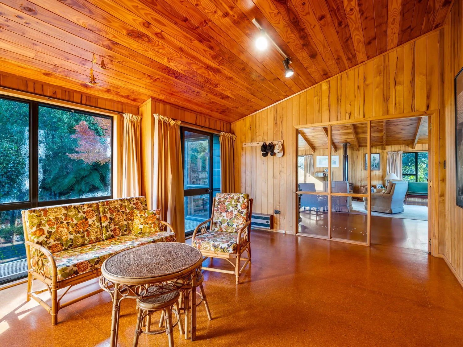 A wooden interior room with floral cushioned wicker furniture a round wicker table and glass sliding doors at Lake View Lodge - Taupo Holiday Home in Pukawa Bay