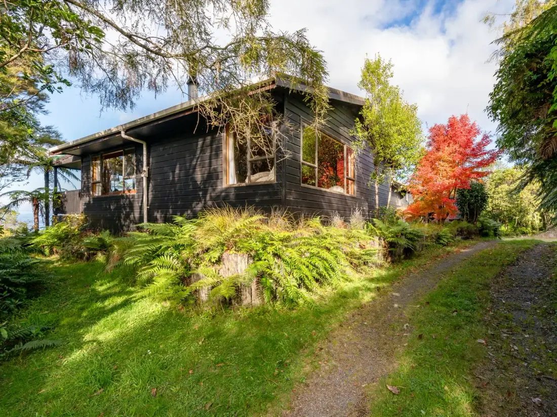 A black wooden house surrounded by green grass trees and a dirt pathway at Lake View Lodge Taupo Holiday Home in Pukawa Bay