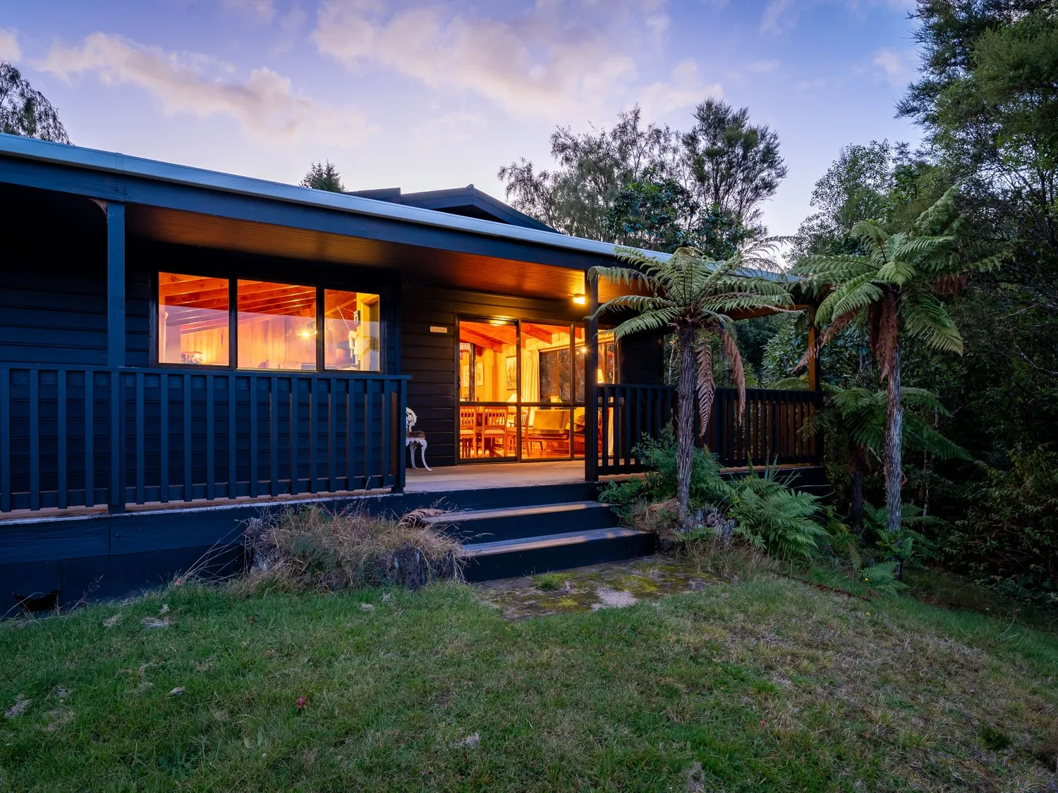 A dark house exterior with warmly lit windows, a porch, and trees at dusk.