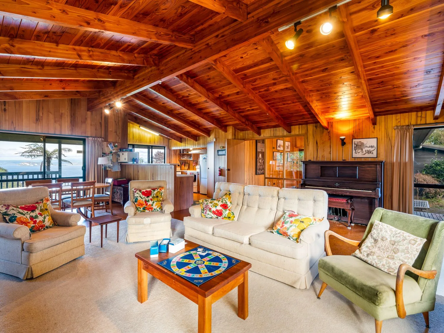 A living room with a beige sofa and armchairs a green armchair a wooden coffee table with a board game a dining table with chairs a piano and wooden ceiling beams at Lake View Lodge - Taupo Holiday Home in Pukawa Bay