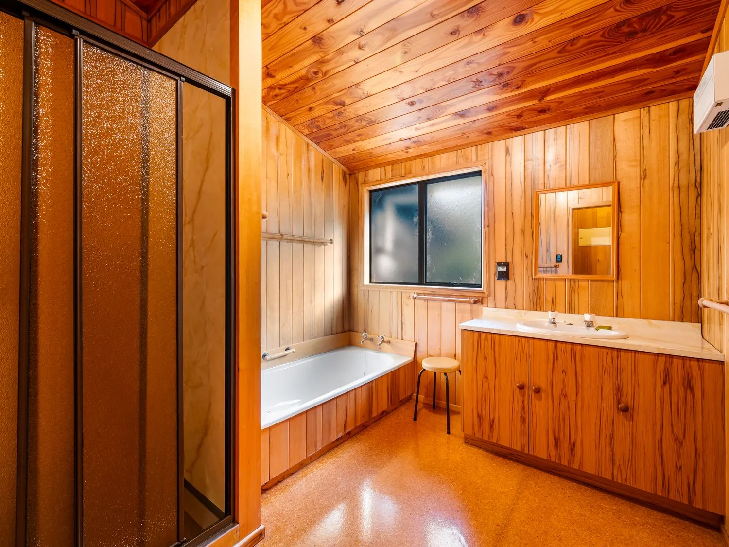 A wood-paneled bathroom with a bathtub, shower, vanity, and small stool.