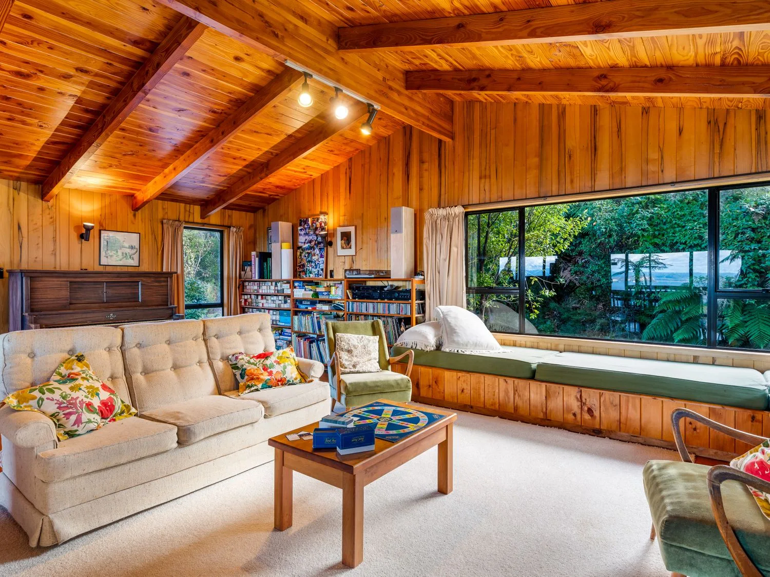 A wood-paneled living room with a beige sofa, bookshelves, and large windows overlooking greenery.